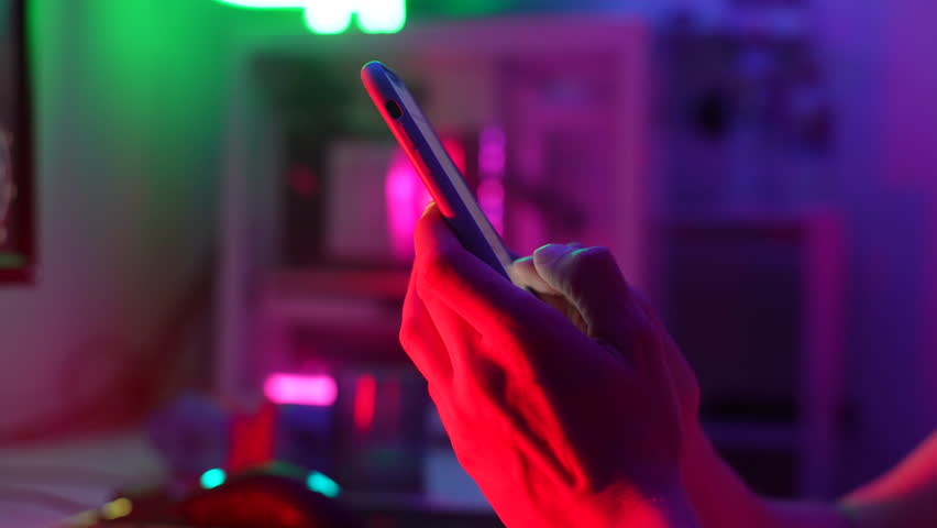 Close-up cropped shot of unrecognizable young man using typing smartphone on dark room with neon bokeh lights. Closeup male hands typing text or shopping online, reading social media or internet. | Shutterstock HD Video #1108649353