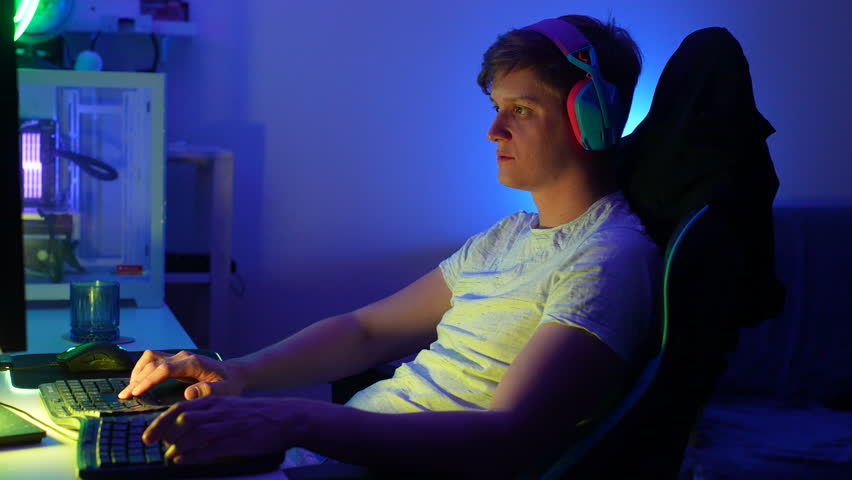 Side view of happy professional male gamer wearing gaming headset having fun playing online video games on personal desktop computer in dark room with neon led lights. Shooting in slow motion.
