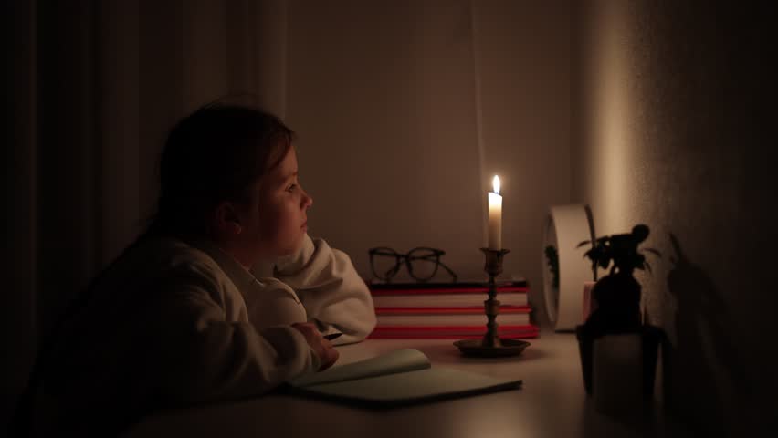 Schoolgirl distance learning at home in a complete darkness without electric lights. Teen kid uses candlelight to do her school homework during blackout. Energy crisis concept by. Blackout in Ukraine. Royalty-Free Stock Footage #1108652839