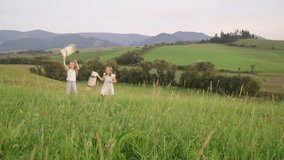 Two sisters running by high green grass meadow with pillows while they have outdoor natural walking, falling down on ground and fooling around. Family values, traveling 4K footage video concept.
