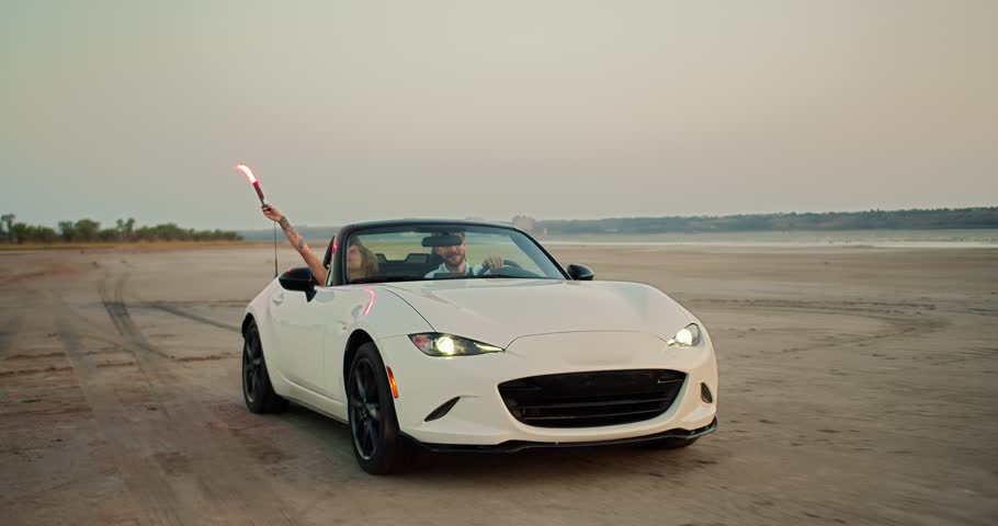 A blonde girl holds hand fire above her head and drive with her bearded guy in a blue shirt on a white convertible car against the river and a gray sky