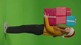 Green screen. Vertical Video Attractive Teenage Girl In Santa Claus Hat Standing Wearing Smiles And Holds A Bunch Of Gift Boxes In Her Hands. Embodying The Spirit Of Celebration, She Radiates Joy