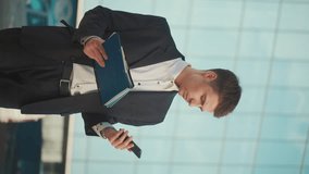 Vertical Video. A Man Engineer or Architect in a Formal Suit and Talking on the Phone Standing Near a Building With Blue Windows and Holding a Blueprint of the Structure and Documents