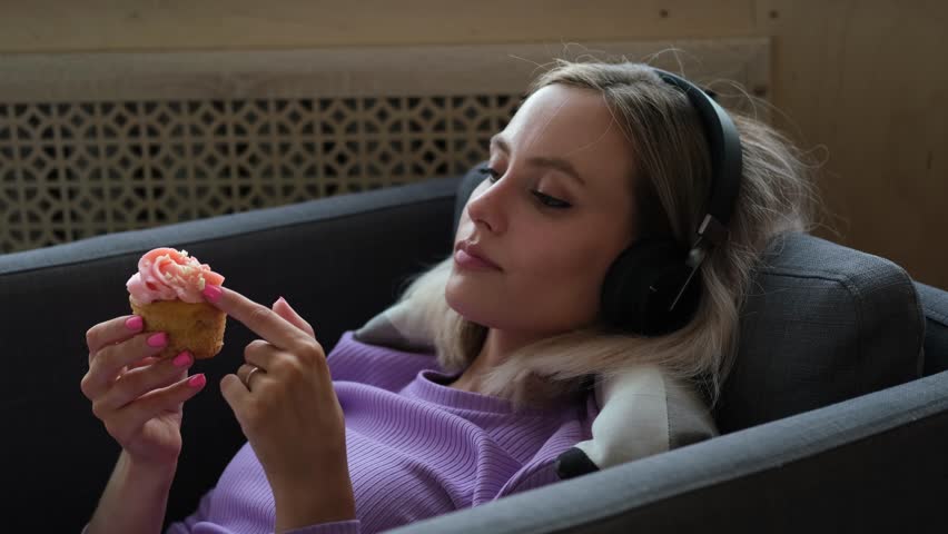 Beautiful sad girl spends time alone at home. The girl listens to music in headphones and eats sweet cakes.