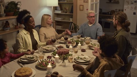 Multi-ethnic family holding each others hands and praying while sitting at festive table during Thanksgiving celebration at home Video de stock