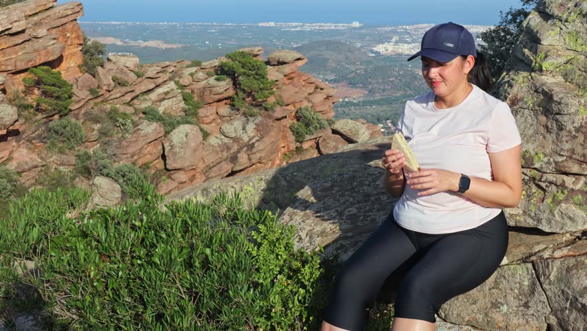 Shot of a young girl on a nature walk pausing to eat sandwich while sitting on a large stone. Rest after climbing the mountain.