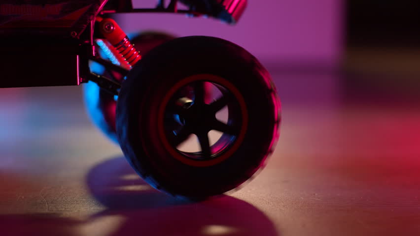Closeup of radio-controlled toy car driving, tugging, spinning wheels on floor in dark living room on background of neon lights. Concept of children toys. Shooting in slow motion.