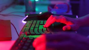 Close-up tracking shot of unrecognizable gamer male pushing keyboard buttons while playing online video game sitting at table with desktop computer, on dark room with neon led lights, slow motion.