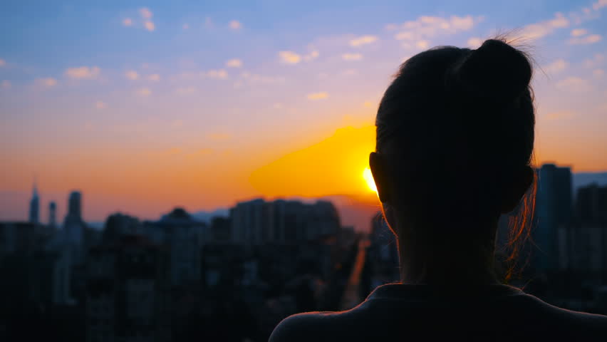 Back view: woman is standing on the balcony and looking at the sunset, sunrise sky over the city - close up, sun lens flares. Lonely, urban, dramatic and freedom concept Royalty-Free Stock Footage #1108696195