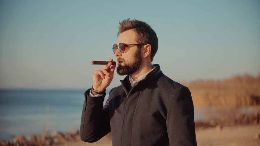 Relaxing Businessman In Sunglasses Smoke Cigar. Stylish Macho In Jacket Taking Break After Hard Business Decision. Confident Well-Dressed Man Smoking. Bearded Man In Sunglasses with  Smoking Cigar