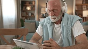 One senior man old caucasian male pensioner sit at home on the sofa bed use digital tablet to watch movie or have a video call online with headphones on his head gray hair and beard