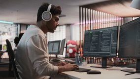 Male Indian Game Programmer Coding On Desktop Computer In Game Development Studio Diverse Office. Focused Man Writes Lines Of Code, Does Gameplay Engineering For New Immersive 3D RPG Video Game.
