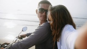 POV of female travel blogger filming vlog, taking selfie photo on smartphone. Young romantic couple sit on motorcycle on sea beach at sunset. Man, woman in love relax on motorbike trip to ocean coast.