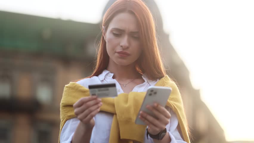 Confused young woman with credit card thoughtfully pondering on screen while has problem with online order payment error or blocked bank account doing shopping at internet on smartphone at modern city Royalty-Free Stock Footage #1108709829