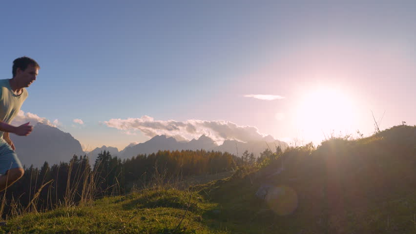CLOSE UP, LENS FLARE: Agile guy runs and jumps with his dog up the trackless road. They are running fast and with light feet to the grassy mountaintop in golden sunset light on a beautiful autumn day.