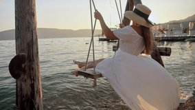 Young woman, with flowing brunette hair in a long white dress, swings in slow motion by the sea beach enjoying sunset. Tourism, the beauty of summertime, holidays, and coastal vacation. - Powered by Shutterstock - Get 15% off with code: PIKWIZARD15