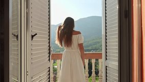 Young brunette woman in a flowing dress opens balcony doors, steps onto the terrace, and revels in a breathtaking mountain sunrise or sunset. Essence of summertime holiday, vacation, nature appreciati - Powered by Shutterstock - Get 15% off with code: PIKWIZARD15