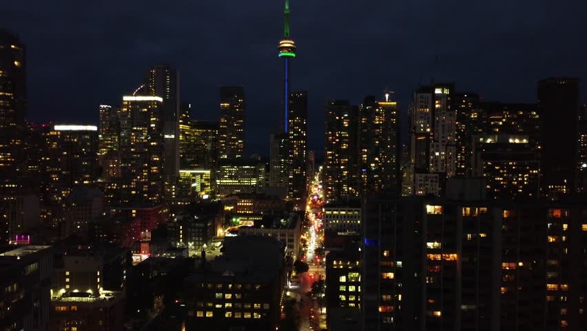 Nighttime rise-up aerial shot of urban downtown Toronto. Drone flying over the financial district and showing the cityscape with the CN tower in the distance. Modern illuminated high-rises.