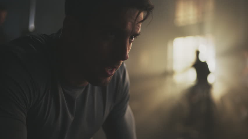 Close-up shot of a tired young man breathing heavily after doing kettelbell lifting exercise while group of people exercising in blurry background