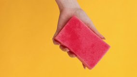 Vertical video. Washing tools. Home hygiene. Woman hand squeezing soft pink household cleaning equipment wet sponge isolated orange background empty space.