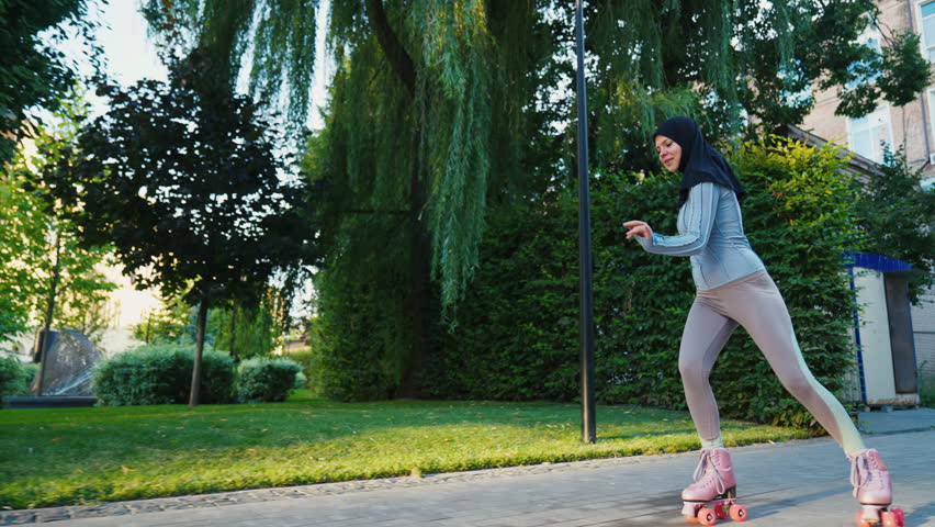 Stable shot of young muslim woman in black hijab riding quad rollers skates in city.
