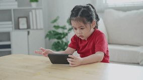 Asian child girl looking using and touch mobile phone screen. Baby smiling funny time to use mobile phone. Too much screen time. Cute girl watching videos while tv in living room, Internet addiction.