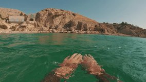 POV shot, man paddling with his hands underwater swims towards rocky shore. First person video, guy swims on the surface of Mediterranean sea, slow motion
