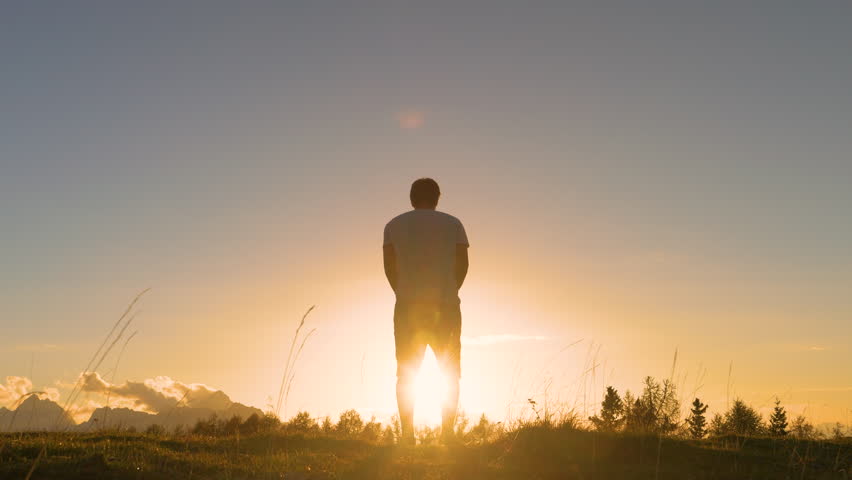 LENS FLARE: Young guy jumps with raised arms on a grassy mountaintop at sunset. He is celebrating as he reaches the top when first rays of autumn morning sun start spilling over the alpine hilltop. Royalty-Free Stock Footage #1108768695