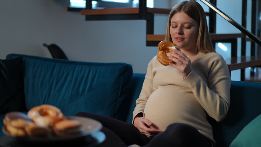 Happy woman caressing pregnant belly biting chewing tasty sweet bun sitting on sofa at home. Portrait of satisfied Caucasian young expectant enjoying delicious pastry dessert indoors