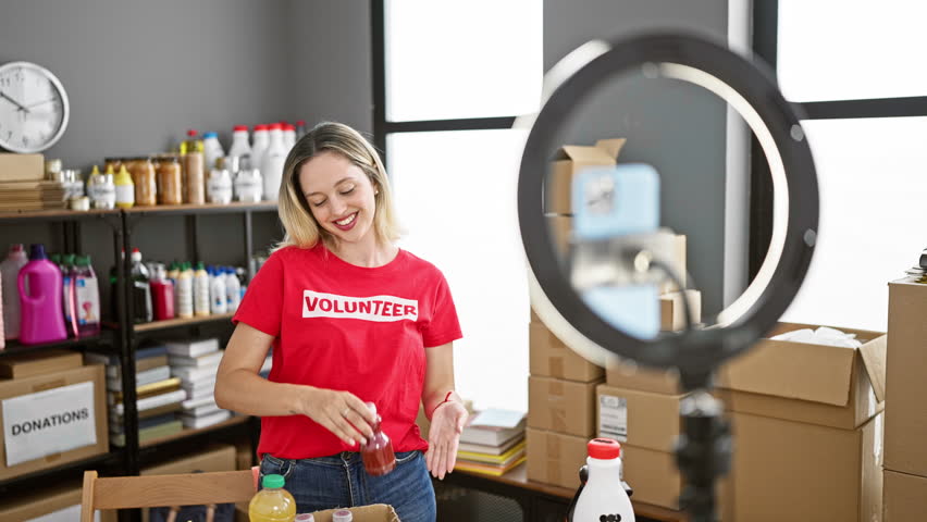 Young blonde woman volunteer showing products to donate recording video at charity center