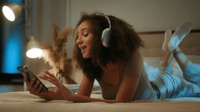 African American ethnic woman lying on cozy bed listen sound in wireless headphones listening music in night evening bedroom relaxed teenage gen z girl with curly hair singing song with mobile phone - Powered by Shutterstock - Get 15% off with code: PIKWIZARD15