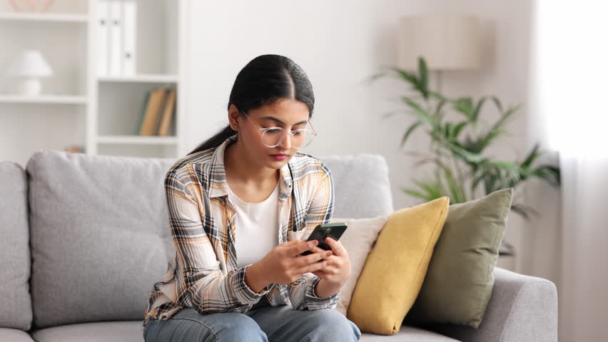 Worried young Indian girl sitting on sofa looking at her phone and does not understand why it is not working. The angry student in glasses does not believe the bad news she saw. Royalty-Free Stock Footage #1108790369