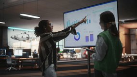 Two Creative Colleagues Collaborating, Pointing at Television Set With Financial Graphs And Charts in Office. Black Female CEO Consults Asian Marketing Manager. Two Women Brainstorming. Slow Motion. - Powered by Shutterstock - Get 15% off with code: PIKWIZARD15