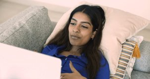 Attractive Indian woman resting on sofa with laptop, smile having pleasant virtual conversation through videocall application, share news and plans on weekend leisure, lead talk lying on couch at home