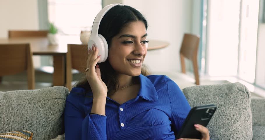 Close up carefree Indian woman listen music through modern wireless headphones holds phone use digital streaming online services enjoy playlist with favourite popular songs spend leisure relax on sofa Royalty-Free Stock Footage #1108798179