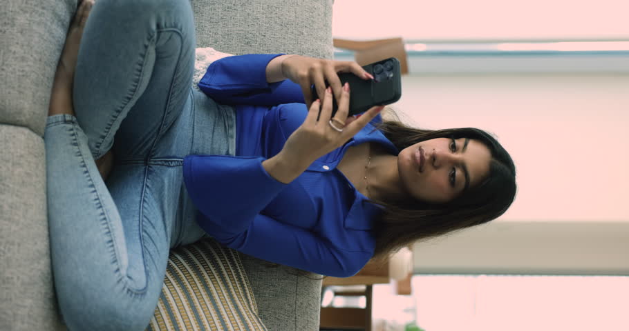 Young Indian woman use cellphone seated on sofa in living room, vertical view. Single satisfied female enjoys new mobile e-dating application, share messages, chat with friend in social media networks Royalty-Free Stock Footage #1108798183