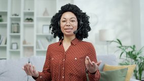 Webcam view. Confident african american businesswoman in headset talking on video call while sitting in home office. Black woman coach conducts an online training, seminar, webinar looking in camera