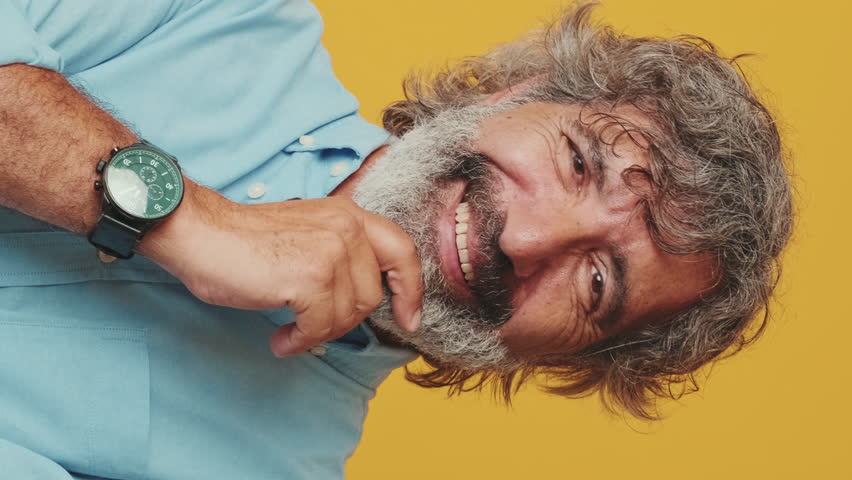 Vertical video, Close-up of an elderly gray-haired bearded man wearing a blue shirt, looking camera, thinking pleasant, smiling, isolated on an orange background in the studio | Shutterstock HD Video #1108808773