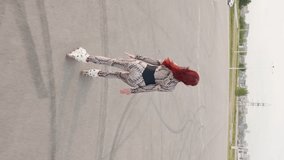 Vertical Video, Slim Woman with Red Hair Has Fun Rollerblading on an Asphalt Parking Lot. Happy Girl Roller-Skates and Raises Her Arms to the Sides, Feeling Freedom and Wellbeing. Slow Motion.