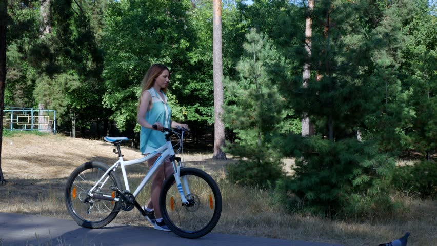 A girl rolls a mountain bike along a park path, and a pigeon runs nearby.