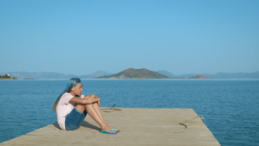 Sea depression. A sad teenage girl sits in a closed pose on a pier against the backdrop of the sea.