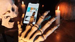 Skeleton using and scrolls smartphone for social networks, in blurred background candles and pumpkin, close up. Horror-loving friends to scariest Halloween party. 