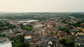 A drone records Heckmondwike, UK, with industrial buildings, bustling streets, and the old town center on a summer evening.