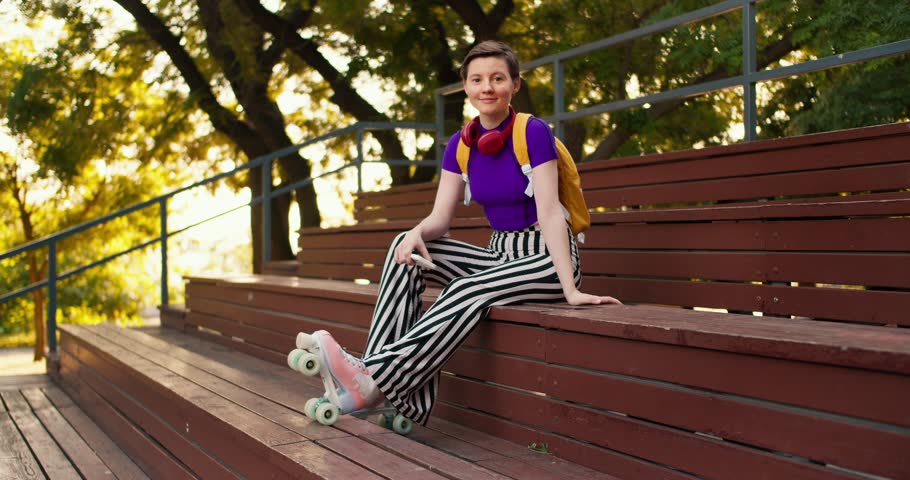 A girl with a short haircut in a purple top, striped pants and a yellow backpack sits on brown benches in the stands in pink roller skates in a skatepark in summer