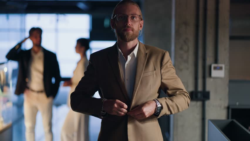 Portrait of a blond man with a beard who adjusts his light brown suit, folds his arms on his chest, smiles and looks at the camera against of a modern office