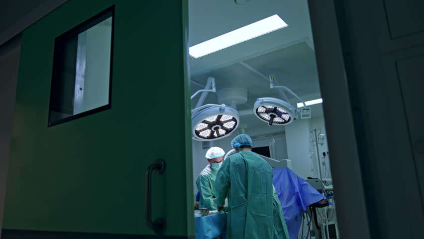 Half opened slider door to a modern surgery room with operation in progress. Three surgeons work hard at the operational table. Royalty-Free Stock Footage #1108835857