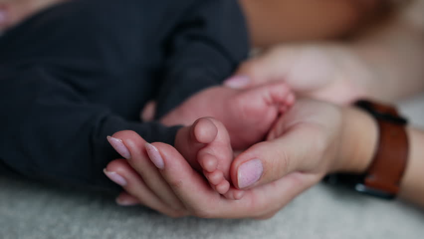 Female hand with brown wrist watch strokes tiny baby toes. Cute infant