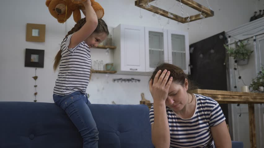 Irritated girl with sore head. Daughter screams and plays with teddy bear. Loud screams annoy sick mother in quarantine. Screaming of children disturbs parent. Mom with sore head is in quarantine. Royalty-Free Stock Footage #1108836055