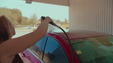 Handheld footage of a woman washing a car at a car wash, using a high-pressure power washerの動画素材