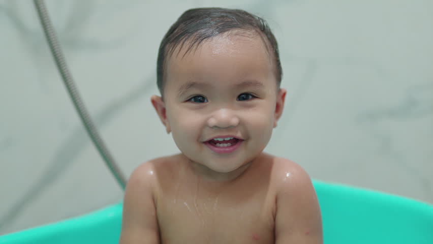 Asian baby girl bathes in a bath with foam and soap bubbles in bathroom.